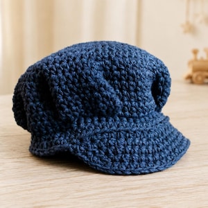 May include: A dark blue newsboy cap crafted from thick, textured yarn. The hat features a rounded, slouchy crown and a short, stiffened brim extending from the front. The crochet construction creates a visible, repeating stitch pattern throughout.