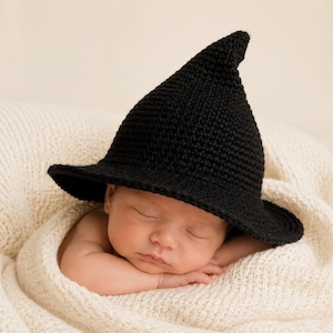 May include: A sleeping baby wearing a black crocheted witch hat, resting on a cream-colored knitted blanket. The hat has a wide brim and a pointed crown. The baby is swaddled in the soft blanket, creating a cozy and adorable scene.