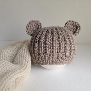 May include: A light brown crocheted hat featuring two small, rounded bear-like ears on top. The hat is constructed with a vertical ribbed stitch pattern and is positioned next to a folded, cream-coloured textured fabric piece.