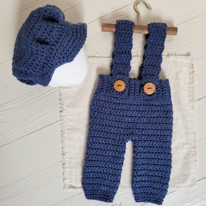 May include: Blue crocheted baby overalls with brown buttons and matching blue crocheted hat.