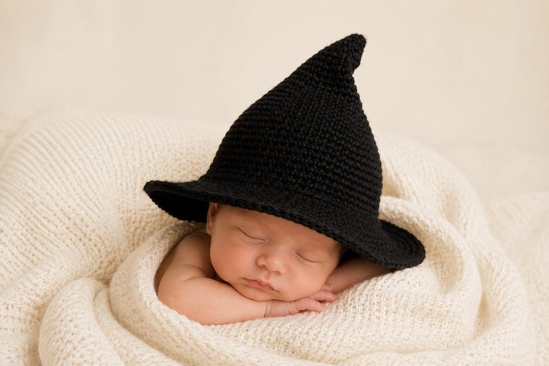 May include: A black crocheted hat with a pointed crown and a wide brim, worn by a sleeping infant wrapped in a soft, cream-colored knitted blanket. The hat features a textured, yarn-based construction with a distinct conical shape and flat brim.
