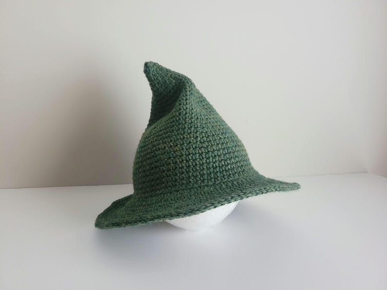 May include: A forest green crocheted hat with a tall, pointed crown and a wide, slightly curved brim. The hat is constructed from a dense, textured yarn stitch pattern, creating a structured yet flexible shape that tapers to a distinct point.