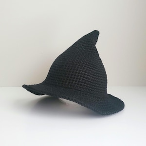 May include: A solid black pointed hat crafted with a textured crochet stitch pattern. The hat features a wide, slightly curved brim and a tall, conical crown that tapers to a rounded point at the top, creating a classic silhouette.