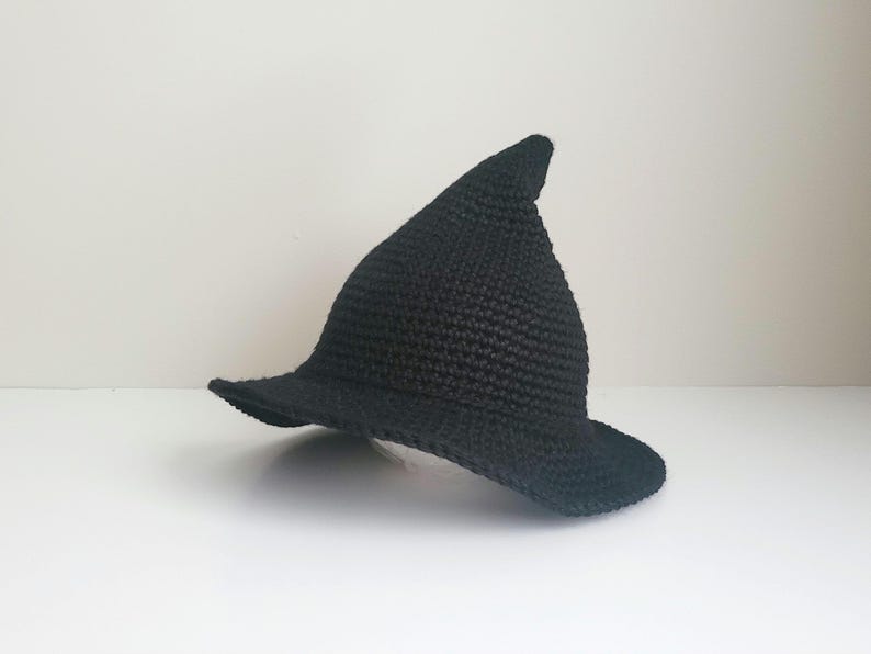 May include: A solid black pointed hat crafted with a textured crochet stitch pattern. The hat features a wide, slightly curved brim and a tall, conical crown that tapers to a rounded point at the top, creating a classic silhouette.