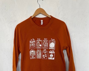 May include: A rust-coloured sweatshirt with a white graphic print of pumpkin varieties. The graphic includes the names "Fairytale", "Atlantic Giant", "Jack Be Little", "Lumina", "Jack Lantern", "Sugar Pie", and "Warty Goblin".