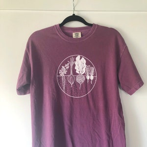May include: A purple t-shirt with a white graphic of a circle with various vegetables inside. The text "COMFORT COLORS" is visible on the tag.