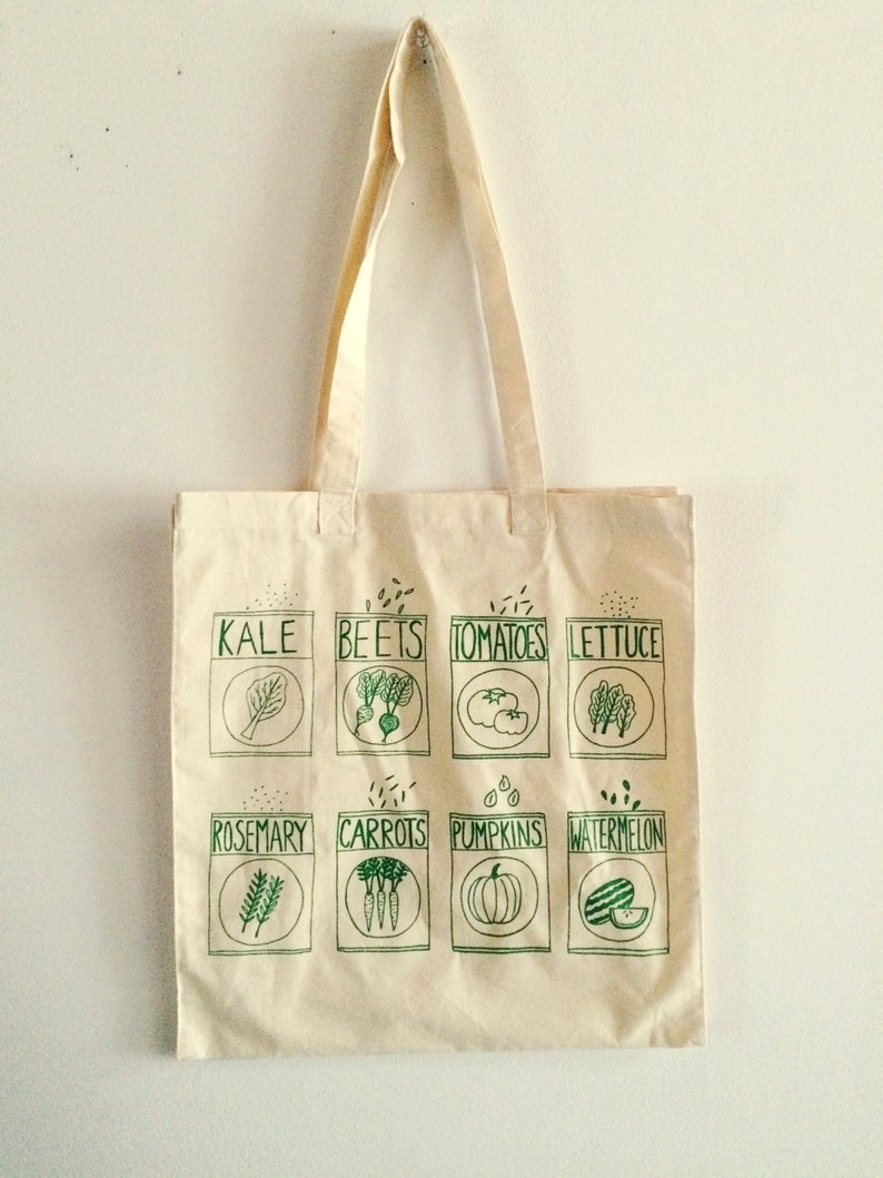 May include: A white canvas tote bag with green text and illustrations of various vegetables and herbs. The text includes the names of the plants: Kale, Beets, Tomatoes, Lettuce, Rosemary, Carrots, Pumpkins, and Watermelon.