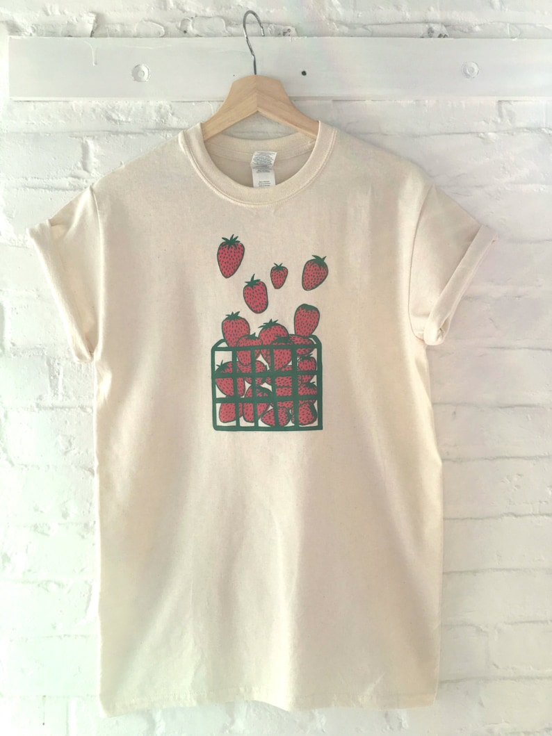 May include: A cream-colored t-shirt with a green and red graphic of strawberries in a basket. The strawberries are spilling out of the basket.