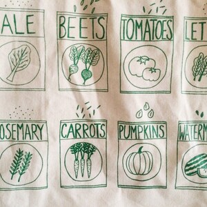 May include: A white tote bag with green illustrations of various vegetables and herbs. The bag features the words "Kale", "Beets", "Tomatoes", "Lettuce", "Rosemary", "Carrots", "Pumpkins", and "Watermelon".