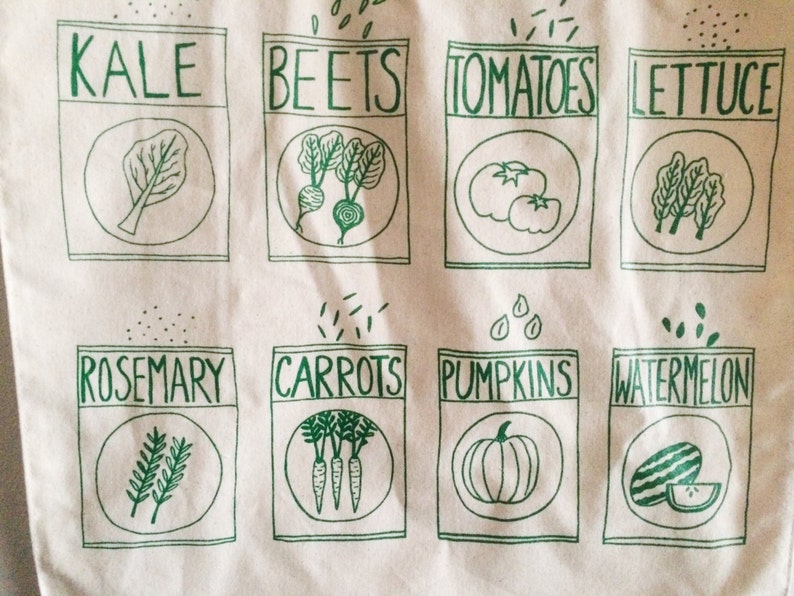 May include: A white tote bag with green illustrations of various vegetables and herbs. The bag features the words "Kale", "Beets", "Tomatoes", "Lettuce", "Rosemary", "Carrots", "Pumpkins", and "Watermelon".