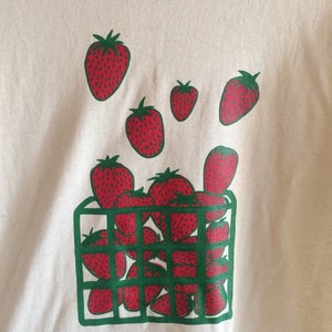 May include: A white t-shirt with a green and red graphic of strawberries in a basket. The strawberries are red with green leaves and are arranged in a basket with a green grid pattern.