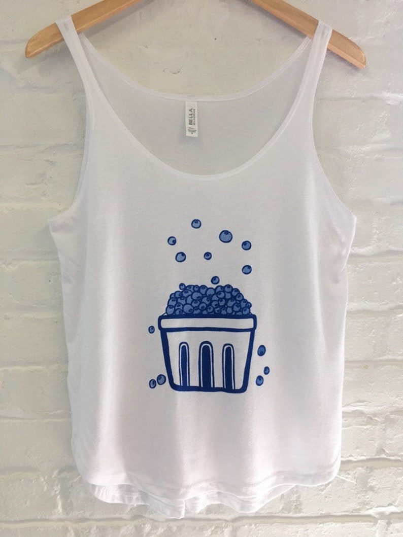 K&ouml;nnte beinhalten: Wei&szlig;es Tanktop mit einer blauen Illustration eines Korbes, der mit Blaubeeren &uuml;berquillt, und Blaubeeren, die um den Korb herum verstreut sind.