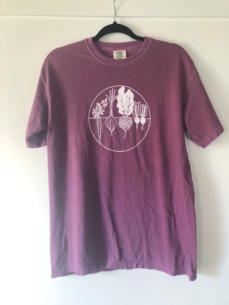 May include: A purple t-shirt with a white graphic of a circle with various vegetables inside. The text "COMFORT COLORS" is visible on the tag.