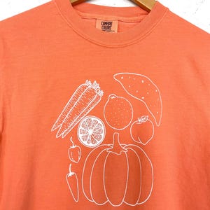 May include: Orange Comfort Colors t-shirt with a white outline illustration of various vegetables and fruits, including carrots, a pumpkin, and an orange slice. The shirt has a crew neck and short sleeves.