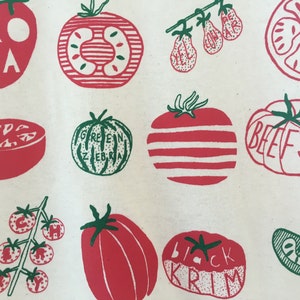 May include: A white canvas tote bag with a red and green illustration of different types of tomatoes. The tomatoes are labeled with their names: Roma, Green Zebra, Beefsteak, Black Krim, and Yellow Pear.