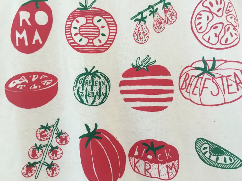 May include: A white canvas tote bag with a red and green illustration of different types of tomatoes. The tomatoes are labeled with their names: Roma, Green Zebra, Beefsteak, Black Krim, and Yellow Pear.