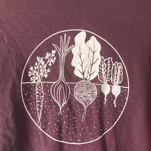 May include: A purple t-shirt with a white graphic of a circle containing a carrot, onion, beet, and two turnips. The text "COMFORT COLORS" is visible on the shirt.