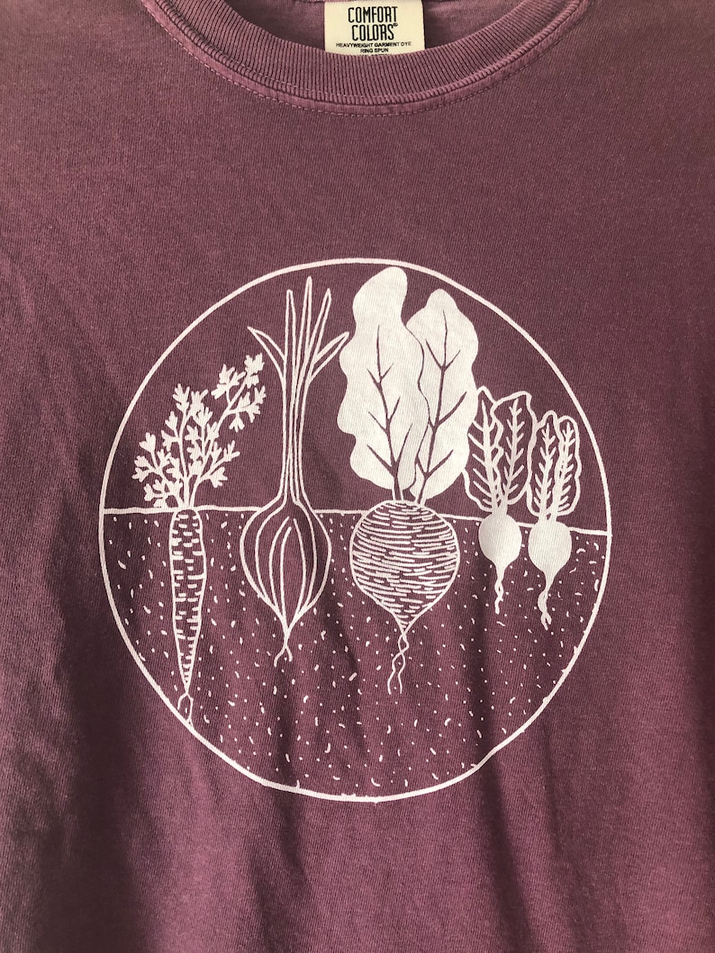 May include: A purple t-shirt with a white graphic of a circle containing a carrot, onion, beet, and two turnips. The text "COMFORT COLORS" is visible on the shirt.
