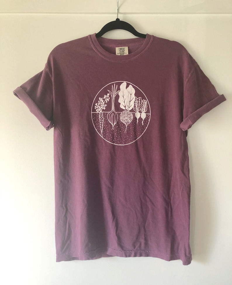 May include: A maroon t-shirt with a white graphic of a circle containing various vegetables, including carrots, onions, radishes, and leafy greens. The text "Comfy Clothes" is visible on the tag.