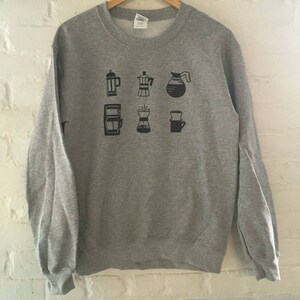 May include: Grey sweatshirt featuring a black graphic of various coffee brewing methods, including a French press, a percolator, a pour-over, and a coffee pot.
