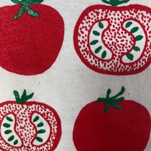 May include: A close-up of a white fabric with a repeating pattern of red tomatoes. Two tomatoes are whole, and two are sliced, revealing the interior. The tomatoes have green stems and are printed in a bold, graphic style.