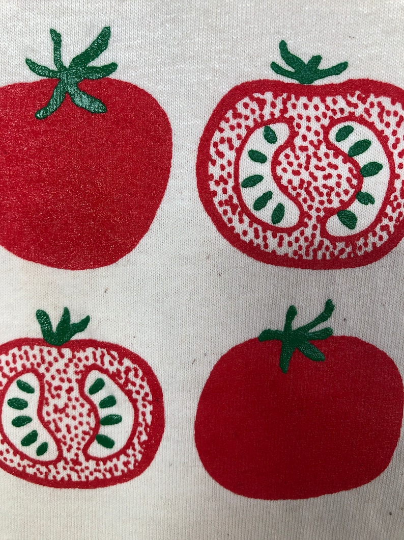 May include: A close-up of a white fabric with a repeating pattern of red tomatoes. Two tomatoes are whole, and two are sliced, revealing the interior. The tomatoes have green stems and are printed in a bold, graphic style.