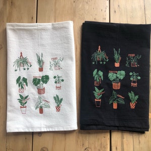 May include: Two kitchen towels with a black and white background. Each towel features a different set of illustrations of various houseplants with their names printed below each plant. The names of the plants include: Pothos, Bird's Nest, Elephant, Pilea, and Spider.