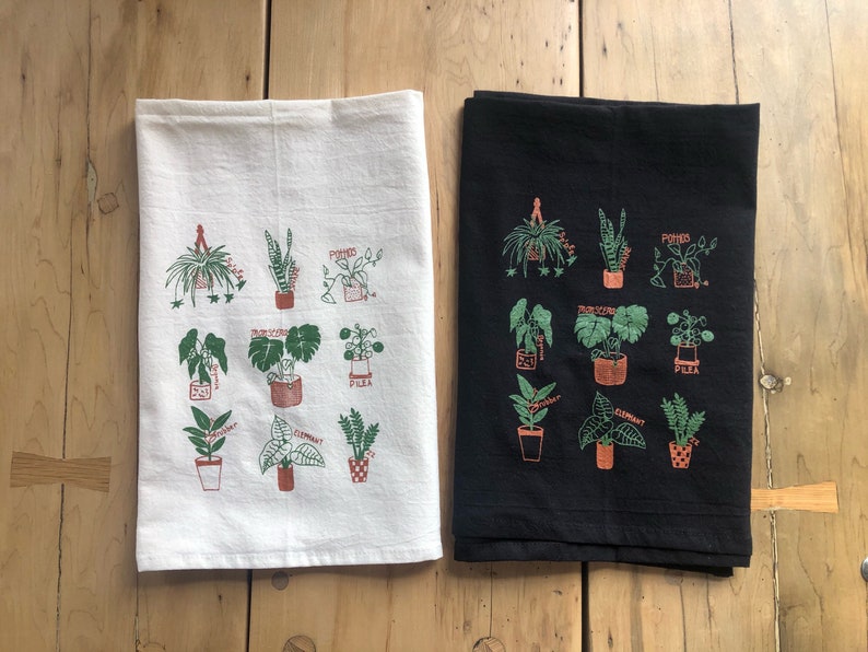 May include: Two kitchen towels with a black and white background. Each towel features a different set of illustrations of various houseplants with their names printed below each plant. The names of the plants include: Pothos, Bird's Nest, Elephant, Pilea, and Spider.