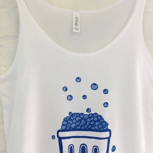 K&ouml;nnte beinhalten: Wei&szlig;es Tanktop mit einer blauen Illustration eines Korbes, der &uuml;berquillt mit Blaubeeren. Blaubeeren sind auch um den Korb herum verstreut.