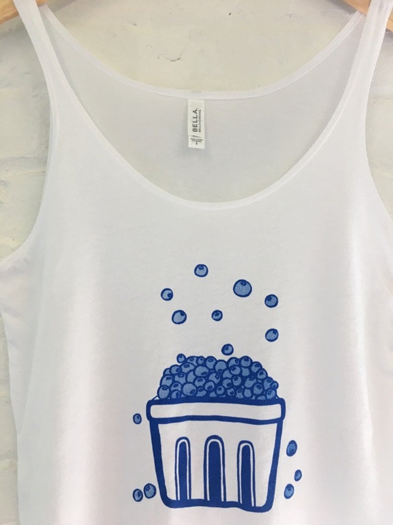 K&ouml;nnte beinhalten: Wei&szlig;es Tanktop mit einer blauen Illustration eines Korbes, der &uuml;berquillt mit Blaubeeren. Blaubeeren sind auch um den Korb herum verstreut.
