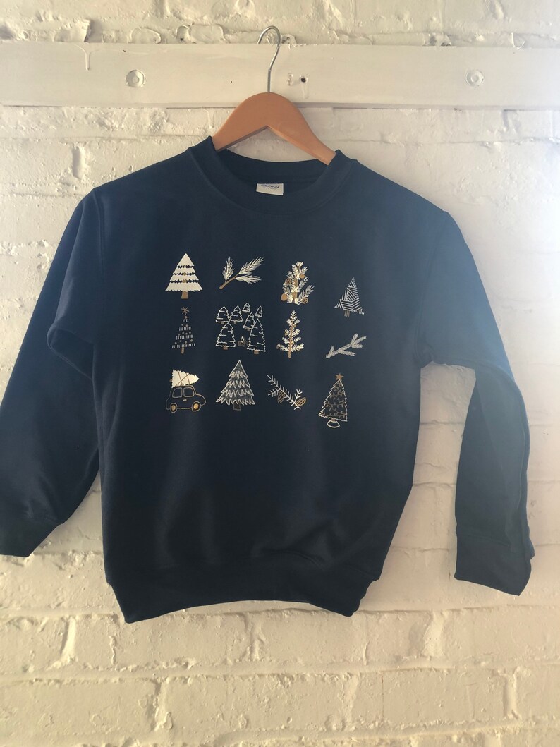 May include: A navy blue sweatshirt with a gold and white design of Christmas trees, branches, and a car carrying a Christmas tree.
