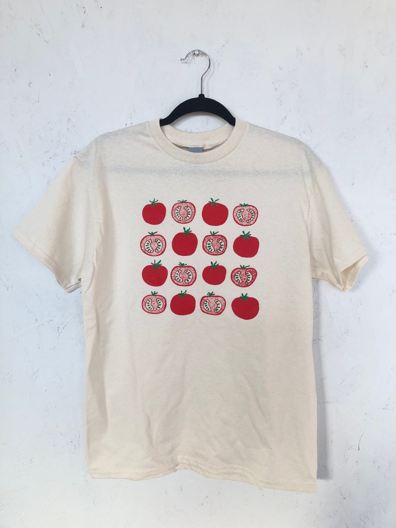 May include: Cream-colored t-shirt featuring a repeating pattern of red tomatoes, some whole and some sliced to reveal the interior. The shirt is hanging on a black hanger against a white wall.