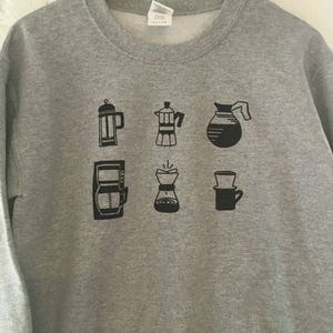 May include: Grey sweatshirt with a black graphic of different coffee brewing methods, including a French press, a percolator, a pour-over, and a drip coffee maker.