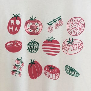 May include: White t-shirt with a graphic print of different types of tomatoes. The tomatoes are illustrated in red and green with names like "Roma", "Beefsteak", "Green Zebra", and "Black Krim".