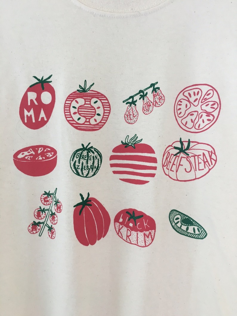 May include: White t-shirt with a graphic print of different types of tomatoes. The tomatoes are illustrated in red and green with names like "Roma", "Beefsteak", "Green Zebra", and "Black Krim".