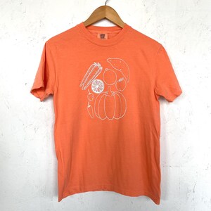 May include: Orange t-shirt with a white outline illustration of various vegetables and fruits, including a pumpkin, sweet potato, and lemon. The shirt is hanging on a wooden hanger against a white wall.