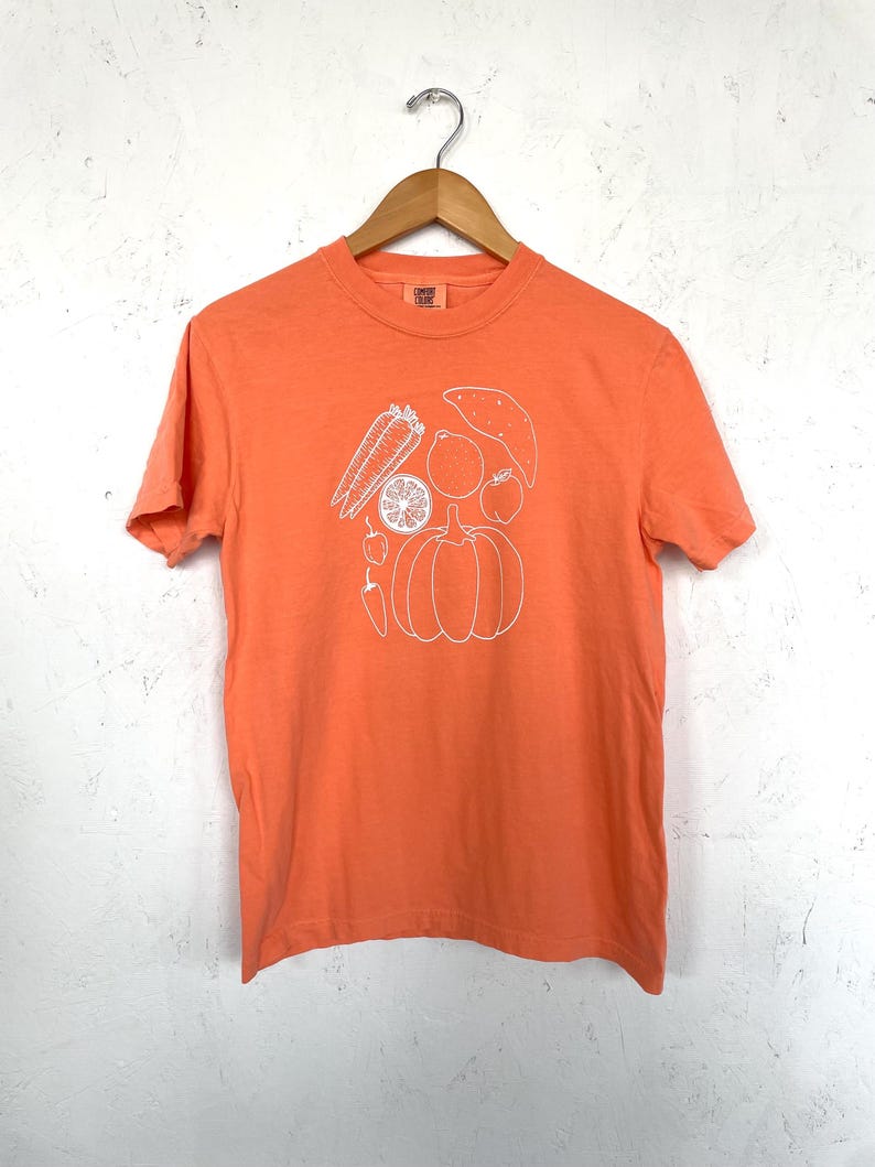 May include: Orange t-shirt with a white outline illustration of various vegetables and fruits, including a pumpkin, sweet potato, and lemon. The shirt is hanging on a wooden hanger against a white wall.