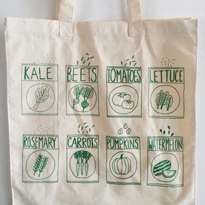 May include: A white canvas tote bag with green illustrations of various vegetables and herbs. The bag has a green print of the words "Kale", "Beets", "Tomatoes", "Lettuce", "Rosemary", "Carrots", "Pumpkins", and "Watermelon".