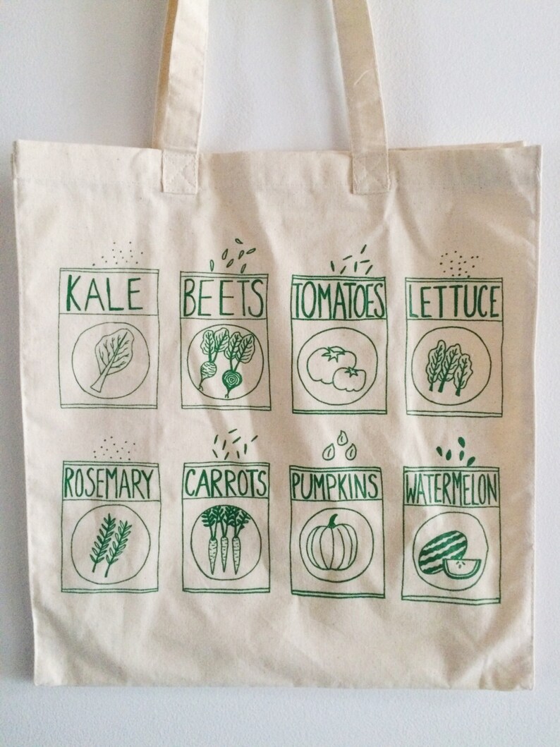 May include: A white canvas tote bag with green illustrations of various vegetables and herbs. The bag has a green print of the words "Kale", "Beets", "Tomatoes", "Lettuce", "Rosemary", "Carrots", "Pumpkins", and "Watermelon".