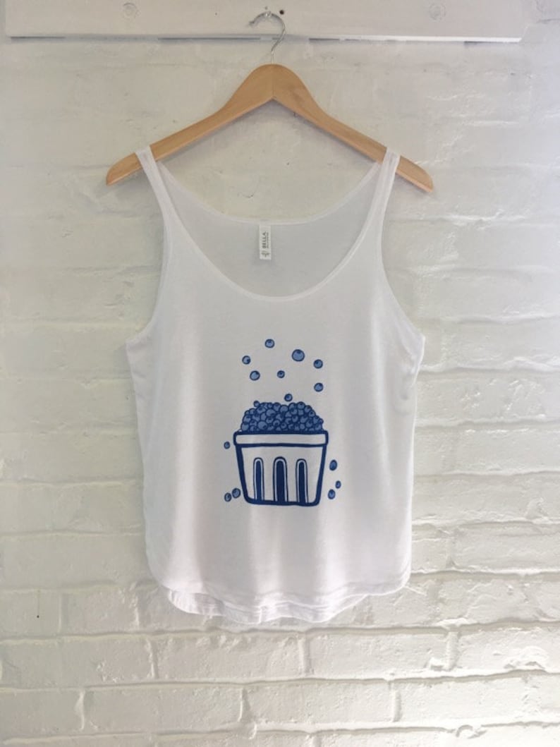 K&ouml;nnte beinhalten: Wei&szlig;es Tanktop mit einer blauen Illustration eines Korbes, der &uuml;berquillt mit Blaubeeren. Die Blaubeeren sind um den Korb herum verstreut.