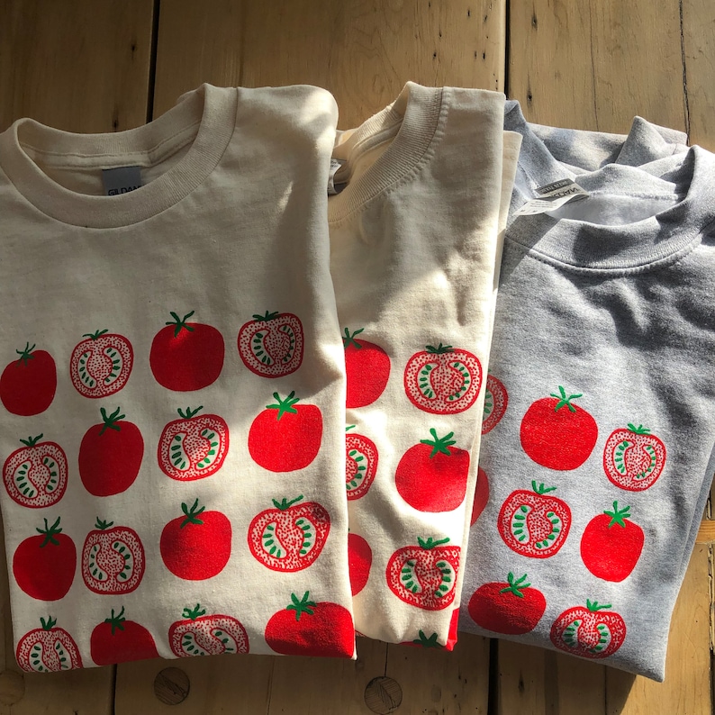 May include: Three t-shirts with a tomato design. The shirts are in cream and gray. The tomato design is red with green stems and leaves. Some tomatoes are whole, and some are sliced to show the inside.