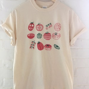 May include: A cream-colored t-shirt with a graphic print of various red tomatoes with green stems and leaves. The tomato varieties are labeled with names like "Roma", "Beefsteak", and "Black Krim".