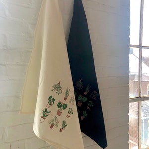 May include: Two kitchen towels hanging on a hook. One towel is white with a green and brown print of various potted plants. The other towel is black.
