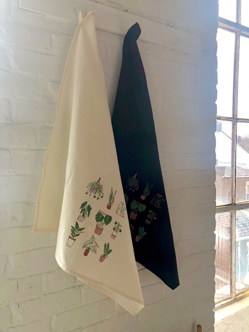 May include: Two kitchen towels hanging on a hook. One towel is white with a green and brown print of various potted plants. The other towel is black.