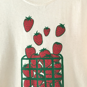 May include: A white t-shirt with a green and red graphic print of strawberries in a basket. The strawberries are red with green leaves and are arranged in a basket with green bars.