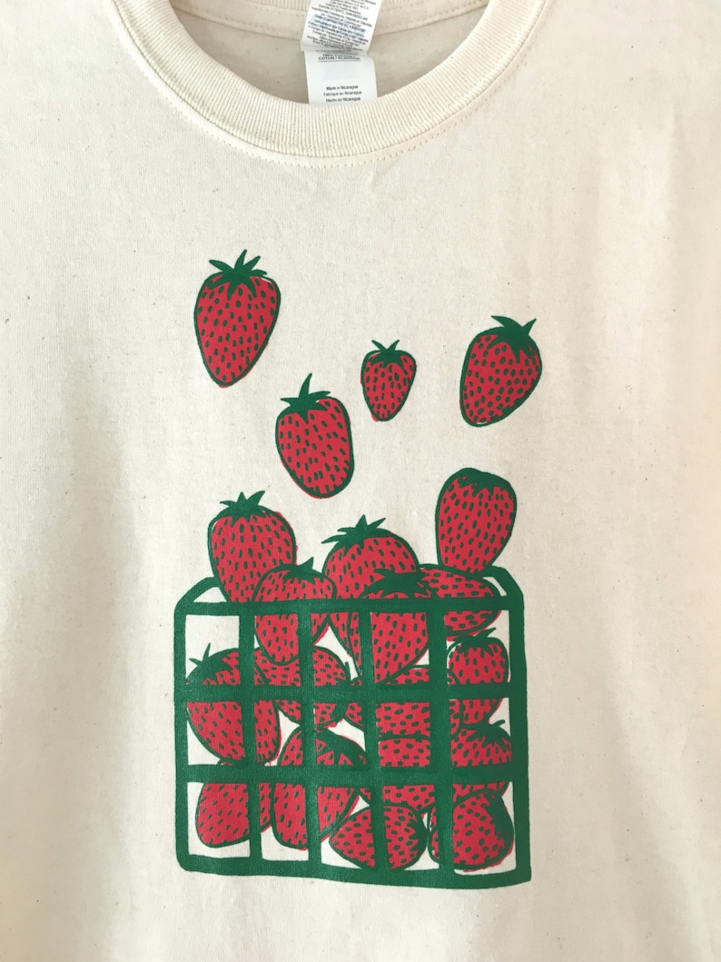 May include: A white t-shirt with a green and red graphic print of strawberries in a basket. The strawberries are red with green leaves and are arranged in a basket with green bars.