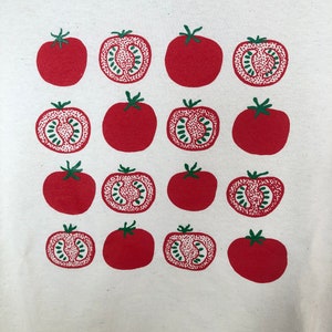 May include: A white t-shirt featuring a repeating pattern of red tomatoes. Some tomatoes are whole, while others are cut in half, revealing a green interior. The tomatoes have green stems.