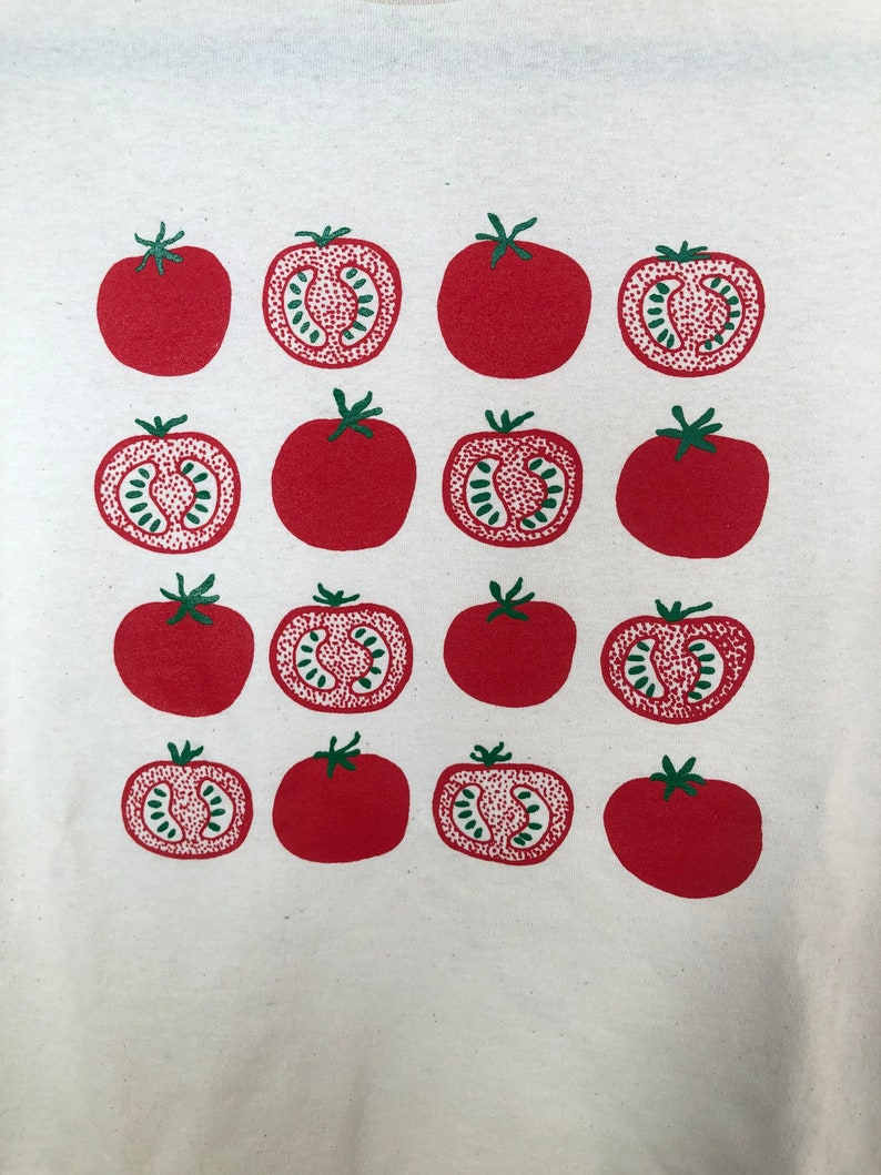 May include: A white t-shirt featuring a repeating pattern of red tomatoes. Some tomatoes are whole, while others are cut in half, revealing a green interior. The tomatoes have green stems.