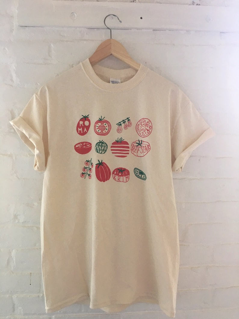 May include: A cream colored t-shirt with a graphic print of red and green tomatoes. The text on the shirt reads "Roma, Beefsteak, Krim".
