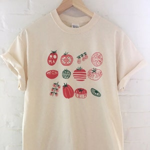 May include: A cream colored t-shirt with a graphic print of various types of tomatoes. The tomatoes are illustrated in red and green with the names of the varieties written on them, including Roma, Beefsteak, and Black Krim.